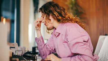 Frau sitzt am Schreibtisch im Büro und hält sich vor Müdigkeit die Hand an den Kopf