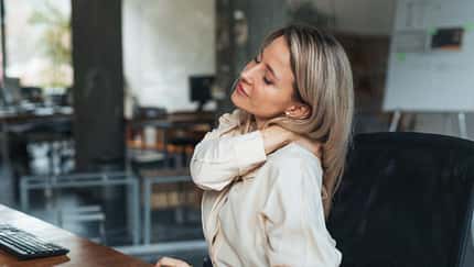 Frau in Businesskleidung sitzt am Schreibtisch und massiert sich den Nacken