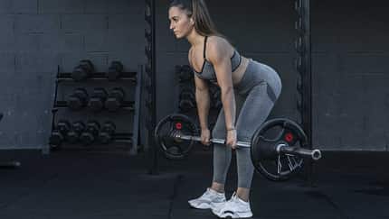 Sportliche Frau führt im Fitnessstudio eine Good Morning Übung mit einer Langhantel aus Sportliche Frau führt im Fitnessstudio eine Good Morning Übung mit einer Langhantel aus