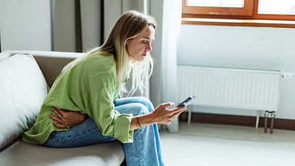 Frau mit Menstruationsbeschwerden sitzt auf dem Sofa und checkt ihre Tracking-App auf dem Smartphone