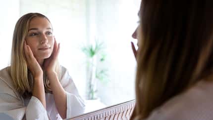 Young blond woman looking at the mirror touching her face. Female examining her facial skin looking in bathroom mirror. Wellness and lifestyle