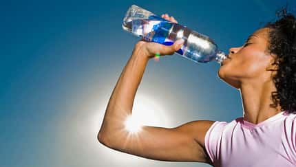 Eine Frau trinkt aus einer Plastikflasche, während im Hintergrund die Sonne scheint