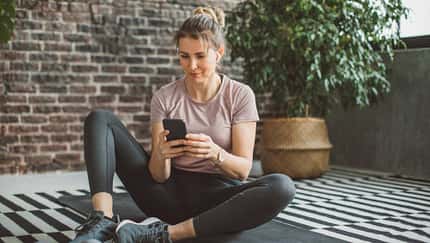 Eine Frau in Trainingskleidung sitzt auf einer Yogamatte und schaut auf ihr Handy Eine Frau in Trainingskleidung sitzt auf einer Yogamatte und schaut auf ihr Handy
