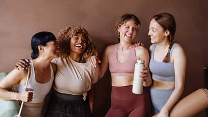 Vier Frauen in Fitnesskleidung stehen lachend vor einem braunen Hintergrund Vier Frauen in Fitnesskleidung stehen lachend vor einem braunen Hintergrund