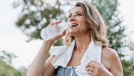 Eine ältere Frau trinkt Wasser nach dem Training mit einem Handtuch über der Schulter