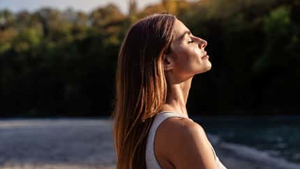 Eine Frau blickt mit geschlossenen Augen in Richtung Himmel. Die Sonne scheint auf ihr Gesicht