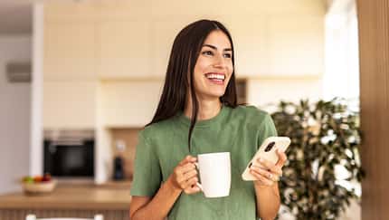 Eine jungeFrau genießt lächelnd ihren Kaffee und hält ihr Handy in der anderen Hand Eine jungeFrau genießt lächelnd ihren Kaffee und hält ihr Handy in der anderen Hand