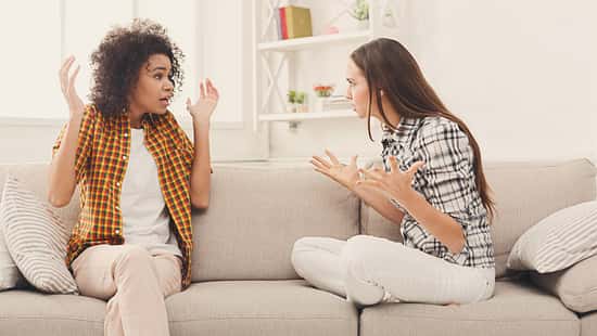 Zwei Frauen sitzen auf einem weißen Sofa und streiten sich. Die linke Frau ist dunkelhäutig, hat braune Locken und trägt ein orangenes Hemd. Die rechte Frau ist weiß, hat braune glatte Haare und trägt ein gemustertes Hemd.