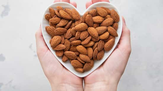 Female hands holding heart shaped plate with peeled almond nuts on light background.Macro.