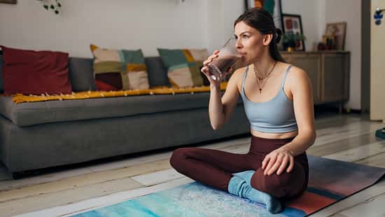 Millennial woman drinks a milkshake while sitting on an exercise mat in the living room