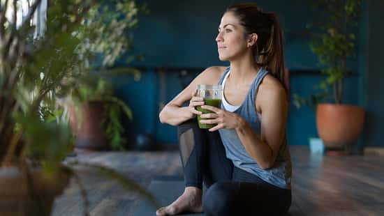 Frau sitzt in Sportkleidung auf einer Yoga-Matte und trinkt grünen Tee