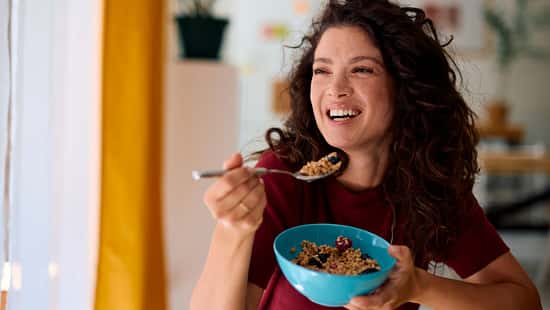 Eine Frau isst lachend ein gesundes Müsli aus einer blauen Schale 