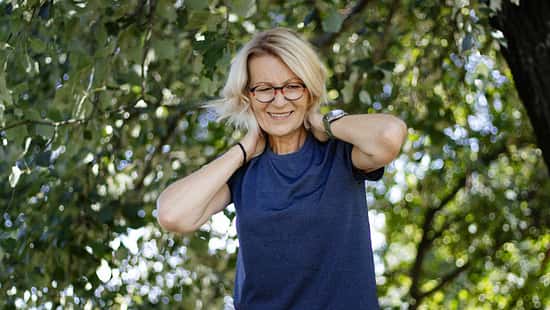 Eine lächelnde ältere Frau mit blonden Haaren und Brille steht im Freien unter grünen Bäumen, trägt ein dunkelblaues T-Shirt und legt entspannt beide Hände in den Nacken.