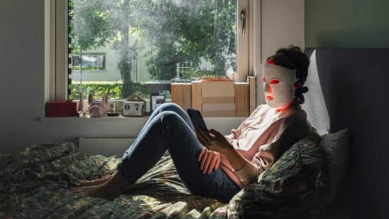 Frau sitzt auf dem Bett am Fenster, trägt eine LED-Maske und schaut auf ihr Smartphone