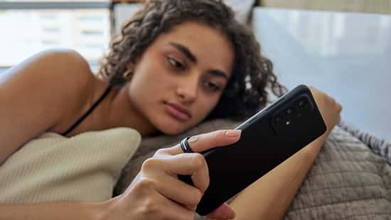 Frau liegt im Bett, blickt auf ihr Smartphone und trägt einen Smart Ring am Finger.