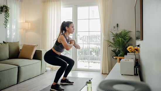 Frau in Sportkleidung macht Kniebeugen auf einer Matte im Wohnzimmer vor einer Terrassentür, neben Sofa und Zimmerpflanze.