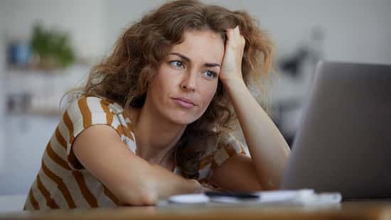 Frau mit lockigen Haaren sitzt nachdenklich vor einem Laptop und stützt den Kopf in die Hand