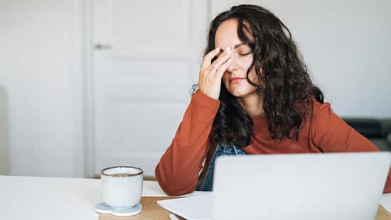 Frau sitzt erschöpft vor einem Laptop, hält sich müde die Stirn, neben ihr stehen eine Tasse und Notizen.