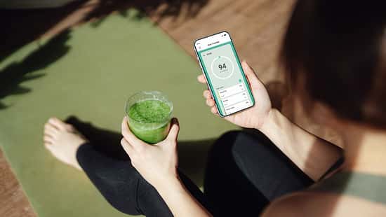 Frau sitzt auf einer Yogamatte, hält einen grünen Smoothie und checkt eine Ernährungs-App auf dem Smartphone.