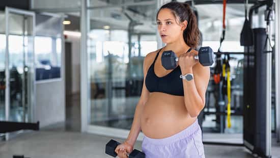 Schwangere Frau steht in einem Fitnessstudio und trainiert mit zwei Hanteln