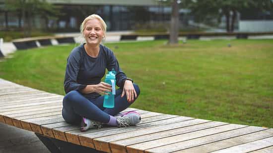 Eine Frau sitzt in Sportkleidung auf einer Parkbank und hält eine Wasserflasche in ihrer Hand