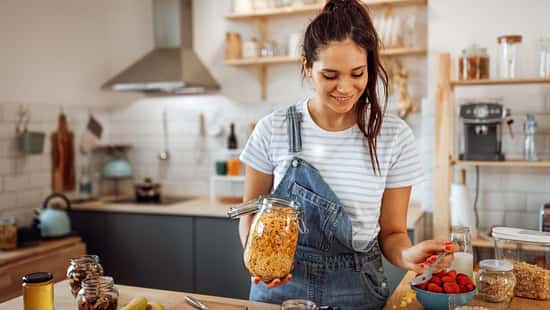 Eine junge Frau in Latzhose steht lächelnd in ihrer Küche und bereitet sich ein Müsli vor