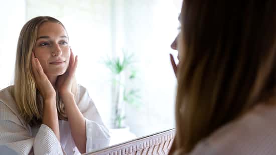 Young blond woman looking at the mirror touching her face. Female examining her facial skin looking in bathroom mirror. Wellness and lifestyle