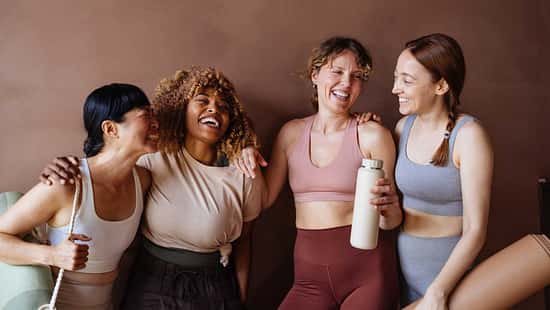 Vier Frauen in Fitnesskleidung stehen lachend vor einem braunen Hintergrund