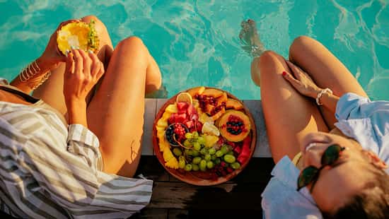 Zwei Frauen sitzen am Pool und essen einen Obstteller
