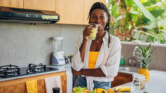 Eine junge Frau steht in ihrer Küche und trinkt einen Smoothie.