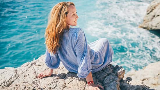 Eine junge Frau ist im Urlaub und schaut auf einem Felsen sitzend zum Meer.