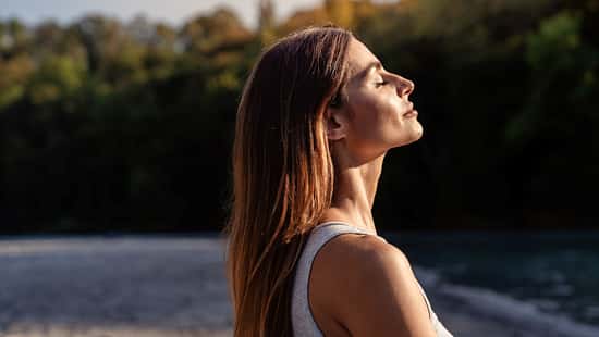 Eine Frau blickt mit geschlossenen Augen in Richtung Himmel. Die Sonne scheint auf ihr Gesicht
