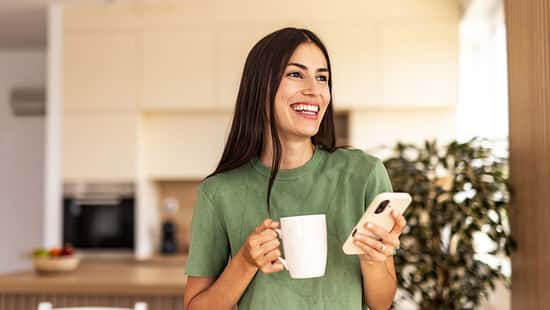 Eine jungeFrau genießt lächelnd ihren Kaffee und hält ihr Handy in der anderen Hand