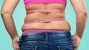 Frau mit sichtbarem Bauchspeck über der Hose wird von hinten mit einem Maßband über dem bauch gezeigt.