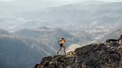 Eine Frau in Laufkleidung läuft einen Bergpass bergauf. Im Hintergrund erstreckt sich ein beeindruckendes Bergpanorama.