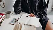 Notizbuch liegt auf Tisch und Frau hält Stift in der Hand.
