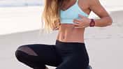 junge, sportliche Frau hockt im Sportoutfit am Strand und schaut sich auf ihren flachen, trainierten Bauch