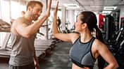 junge, trainierte Frau klatscht mit jungem, trainiertem Mann ein, während die beiden ihr Krafttraining im Fitnessstudio absolvieren