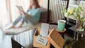 Frau sitzt auf dem Sofa im Hintergrund im Hintergrund. Im Vordergrund steht eine Kiste wo das Handy drin ist, mit einem Zettel dran wo "Digital Detox" draufsteht