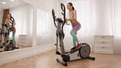 Woman,With,Towel,Using,Modern,Elliptical,Machine,At,Home