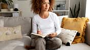 Pensive Young Hispanic woman thinking before writing on personal diary sitting on the couch at home. Multiracial latina female writing notes with pen.