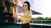 Cheerful caucasian female jogger in headphones enjoying music in headphones and using wearable device on wrist for GPS and time tracking on training, smiling 20s sporty woman looking at smartwatch