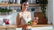 trainierte Frau, die eine gesunde Poke Bowl isst, während sie zu Hause in der Küche in die Kamera schaut