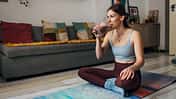 Millennial woman drinks a milkshake while sitting on an exercise mat in the living room