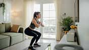 Frau in Sportkleidung macht Kniebeugen auf einer Matte im Wohnzimmer vor einer Terrassentür, neben Sofa und Zimmerpflanze.