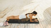 Frau macht draußen eine Unterarmstütz-Plank auf einer Yogamatte auf einem Holzboden.