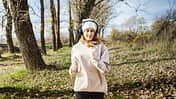Beautiful young woman with wool beanie and wireless headphones jogging and listening to music in nature during cold and sunny autumn day.