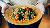Zwei Frauenhände halten eine Schale mit einem Kichererbsen-Spinat-Curry Zwei Frauenhände halten eine Schale mit einem Kichererbsen-Spinat-Curry