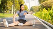 Eine Frau sitzt auf der Straße und stretcht sich in Sportkleidung während sie auf ihre Fitnessuhr schaut