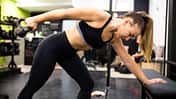 Eine fitte junge Frau macht Trizeps-Kickbacks mit einer Kettlebell gestützt auf eine Bank im Fitnessstudio